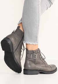 Šedé kožené kotníkové boty s šněrováním vpředu, bočním zipem a kovovým detaily přezky. Texturovaná gumová podrážka pro lepší trakci.