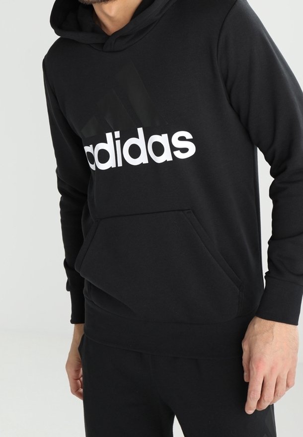 Uomo che indossa una felpa nera Adidas con logo bianco, mani rilassate, mostrato dalle spalle ai fianchi su uno sfondo chiaro uniforme.