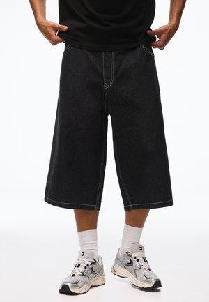 Person trägt weite schwarze Denim-Shorts, weiße Sportsocken und silberne Sportschuhe und steht vor einem schlichten weißen Hintergrund.