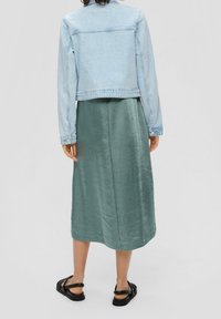 Hellblaue Jeansjacke über einem grünen Satin-Midikleid mit geradem Schnitt und schwarzen Riemchensandalen. Glatte Textur und schlichtes Design.