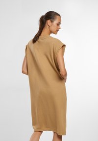Tan oversize bavlněné šaty s krátkými rukávy a kulatým výstřihem. Vyznačují se volným střihem a délkou ke kolenům. Hladká textura.