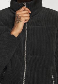 Svart kordflodspuffjacka med hög krage, blixtlåsförsedd framsida och ribbad textur, med elastiska muddar och två fickor med dragkedja.