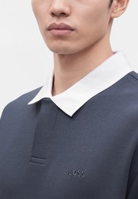 Gros plan d'une personne portant un polo bleu foncé avec un col blanc et le mot BOSS brodé sur la poitrine.