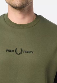Sudadera verde oliva con cuello acanalado y texto negro bordado de "FRED PERRY" con un detalle de corona de laurel debajo del texto.