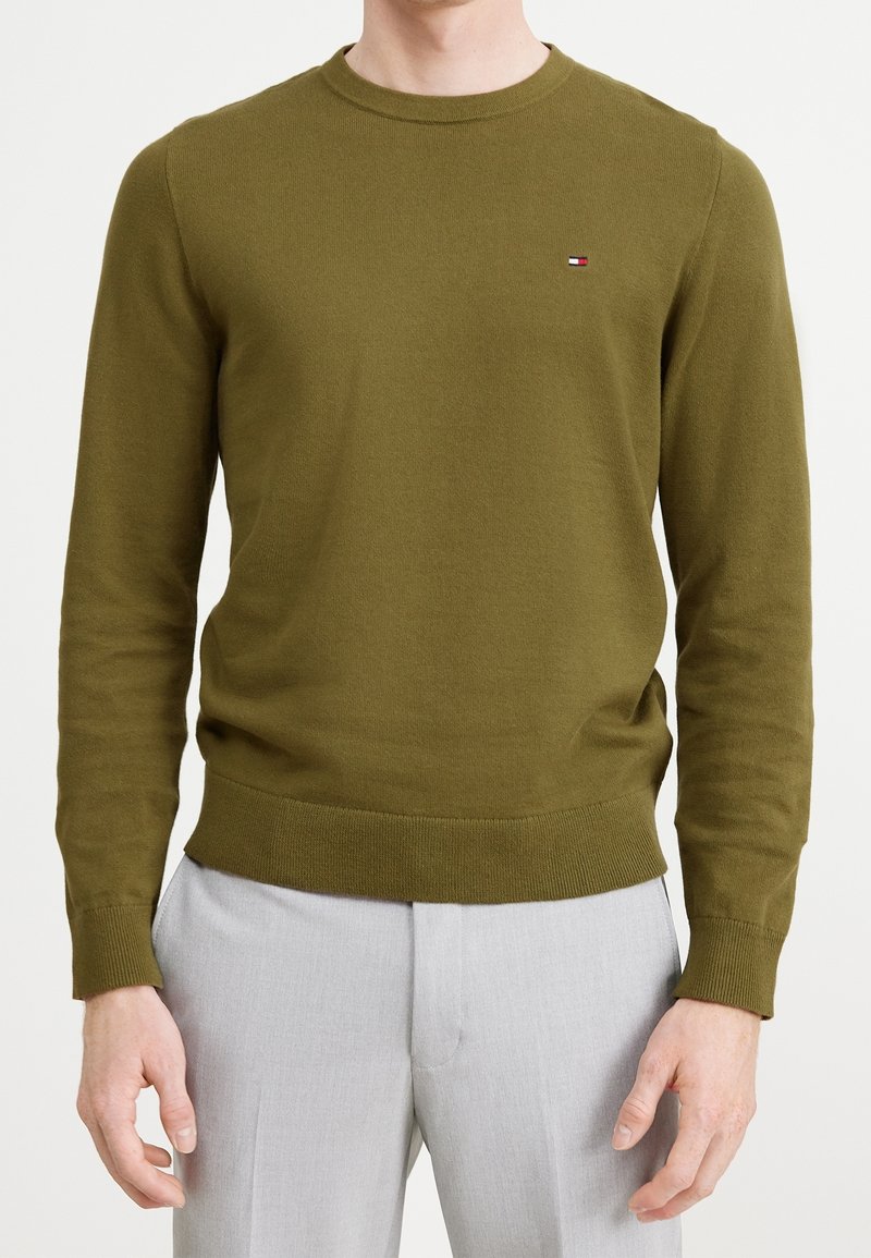 Homme portant un pull col rond vert olive avec un petit logo sur la poitrine et un pantalon gris clair, debout sur fond blanc.
