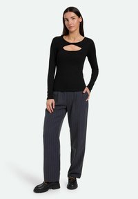 Schwarzes Langarmshirt mit einem Schlüsselaugen-Ausschnitt; kombiniert mit grauen, gestreifen, weiten Hosen und schwarzen Schuhen mit einer klobigen Sohle.