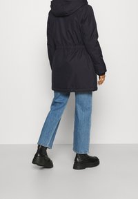 Mörknavy lång jacka med huva, elastisk midja och muddade ärmar, pairad med ljusblå denimjeans och svarta chunky boots.