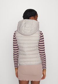 Hellgraue Steppweste mit hohem Kragen und Kapuze, die über ein gestreiftes Langarmshirt getragen wird und horizontale Nähte sowie eine taillierte Passform aufweist.