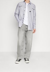 Vaaleansininen pitkähihainen paita, jossa on kaksi rintataskua ja mustat napit, yhdistettynä harmaisiin leveälahkeisiin farkkuihin ja mustiin lenkkareihin.