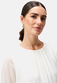 Weiße Bluse mit plissierter Textur, runder Ausschnitt und durchsichtigen langen Ärmeln. Goldene Creolen runden den Look ab.