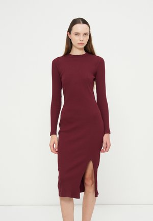 Junge Frau mit langen Haaren, die ein eng anliegendes, geripptes bordeauxfarbenes Kleid mit langen Ärmeln und einem Frontschlitz trägt und vor einem einfarbigen weißen Hintergrund steht.