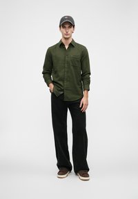 Νεαρός άνδρας που φοράει ένα ελαιόχρωμο πουκάμισο με κουμπιά, μαύρο παντελόνι σε φαρδιά γραμμή, καφέ αθλητικά παπούτσια και γκρι καπέλο baseball, στέκεται με το ένα χέρι στην τσέπη.