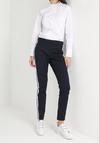 Granatowe spodnie z białymi paskami po bokach, w połączeniu z białą bluzką z długim rękawem i falbaniastym dekoltem. Noszone z białymi trampkami.