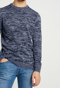 Pull bleu marine avec un motif texturé et moucheté, manches longues et col rond, associé à un jean bleu clair.