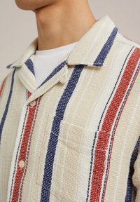 Skjorta med knappar fram i krämfärgad texturerad väv, med röda och blå vertikala ränder och en bröstficka. Krage.