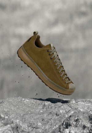 Brauner Wildleder-Sneaker mit beigen Schnürsenkeln in der Luft über felsiger Oberfläche, sichtbare Schmutzpartikel, die von der Sohle fallen.