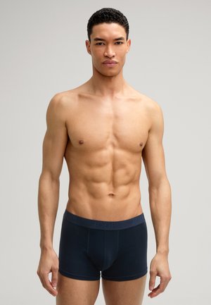 Muskelöser Mann mit kurzen lockigen schwarzen Haaren, der vor einem neutralen Hintergrund steht und dunkelblaue eng anliegende Boxershorts trägt.