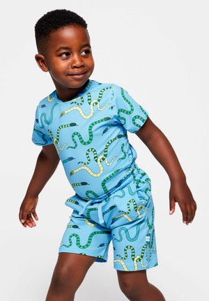 Niño pequeño sonriendo, vestido con camiseta y pantalones cortos a juego de color azul claro con patrones de serpientes verdes y amarillas, de pie frente a un fondo liso.