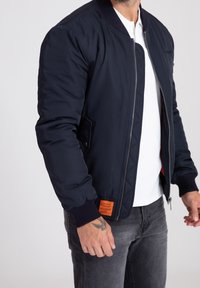 Muž v námořnické bomber bundě s oranžovou nášivkou, bílé polo tričko, šedé džíny a viditelným tetováním na levé ruce na jednobarevném pozadí.