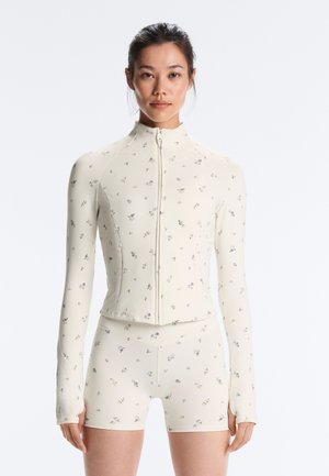 Femme portant une veste zippée ajustée couleur crème assortie à un short avec un petit imprimé floral, debout devant un fond uni.