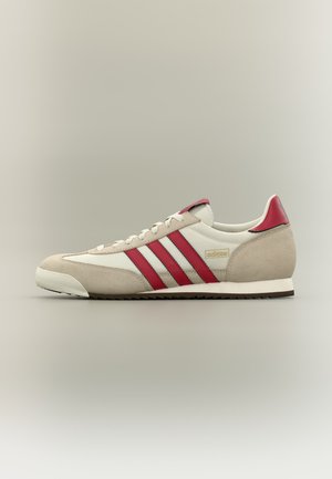 Beige og hvid sneaker med røde striber, rødt hælskinne, ruskindsnæse og hvid sål, set fra ydersiden mod en neutral baggrund.