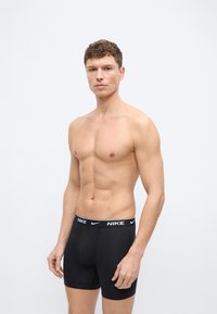 Ανδρας χωρίς πουκάμισο με κοντά σγουρά μαλλιά στέκεται με το πρόσωπο προς τα εμπρός φορώντας μαύρα μποξεράκια Nike με λογότυπο στη ζώνη μέσης.