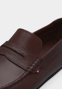 Καφέ δερμάτινο μοκασίνι με ραμμένο λουρί πάνω από τη φτέρνα και μια μικρή υφασμάτινη ετικέτα σε κόκκινο, λευκό και μπλε στη πλευρά.