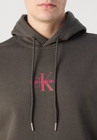 Felpa con cappuccio verde scuro realizzata in tessuto misto cotone. Presenta un logo rosa sul petto e un cappuccio con cordino con estremità in metallo argentato.