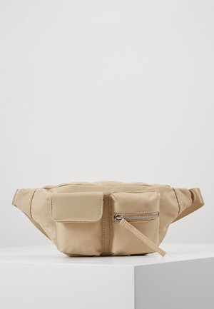 Beige bæltetaske med to forlommer, den ene med klap og den anden med lynlås, hvilende på en hvid overflade mod en ensfarvet baggrund.