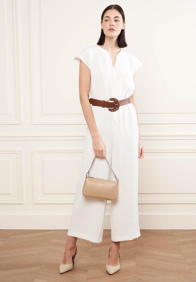 Une combinaison en lin blanc à manches courtes, avec une ceinture marron cintrée avec une boucle ronde, associée à un sac à main beige et des talons de couleur claire.