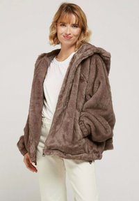 Veste en fausse fourrure couleur brun clair avec une capuche, fermeture éclair et poches latérales. Présente une surface douce et texturée ainsi qu'une coupe ample et décontractée.