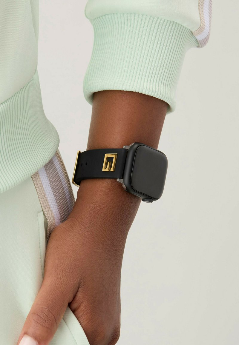 Montre connectée noire avec un cadran carré, accents dorés sur le bracelet, sur un fond en tissu vert clair.