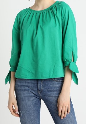 Femme portant un chemisier vert ample à manches trois-quarts nouées aux poignets, associé à un jean bleu en denim sur un fond uni.