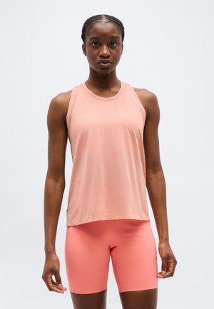 Tânără purtând un top fără mâneci roz deschis și pantaloni scurți mulați roz coral, stând cu fața spre înainte, pe un fundal simplu.