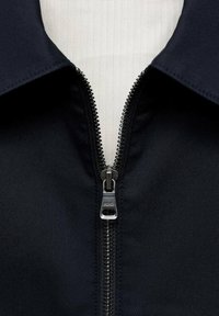 Maritieme jas met een spitse kraag, voorzien van een zilveren rits aan de voorkant, textuurstof en zichtbare stikseldetails.