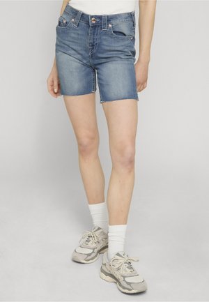 Osoba ubrana w średniobłękitne szorty dżinsowe z surowymi brzegami, białe skarpetki i beżowo-szare sneakersy, stoi na tle jednolitego szarego tła.