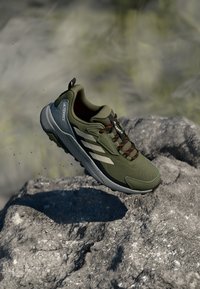 Grüner Trailrunning-Schuh mit einem atmungsaktiven Mesh-Obermaterial, Gummisohle, orangefarbenen Akzenten und drei weißen Streifen an der Seite.