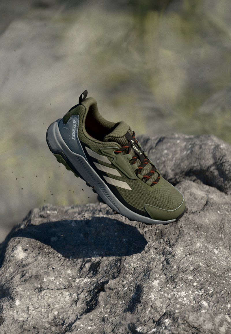 Grüner Trailrunning-Schuh mit einem atmungsaktiven Mesh-Obermaterial, Gummisohle, orangefarbenen Akzenten und drei weißen Streifen an der Seite.