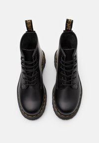 Bottes en cuir noir à lacets avec surpiqûres jaunes, semelles épaisses en caoutchouc et languettes avec inscriptions jaunes, vues de dessus sur fond blanc.