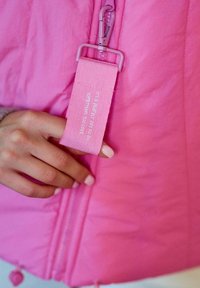 Růžová nadýchaná bunda s předním zipem a textilní cedulkou. Cedulka obsahuje bílý nápis: "Je to perfektní den na to dělat něco, co milujete."