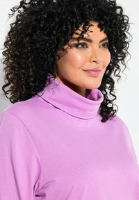 Maglione a collo alto lungo, di un delicato colore lilla, con una texture liscia e una vestibilità comoda. Dettaglio del colletto ripiegato. Capelli naturali e ricci visibili.