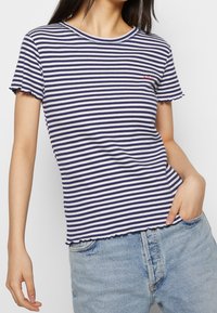 Pruhované tričko s námořnickými a bílými vodorovnými pruhy, ribbovaná textura, kulatý výstřih a volánkové okraje rukávů. Malý vyšívaný detail na hrudi.