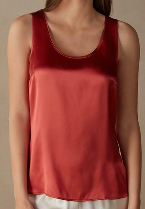 Frau trägt ein ärmelloses, glänzendes rotes Satin-Top, dargestellt von den Schultern bis zur Hüfte vor neutralem Hintergrund.