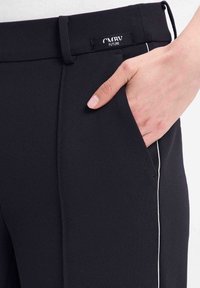 Zwarte op maat gemaakte broek met een textuurstof, voorzien van een zijzak en een witte piping langs de naad. Merktag zichtbaar bij de taille.