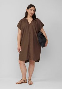 Bruine jurk met korte mouwen en V-hals, van stof met een textuur en een losse pasvorm, gecombineerd met een zwarte geweven tas en bruine sandalen.