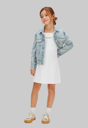 Pige stående iført en lys denimjakke over en hvid Calvin Klein-kjole, med hvide sokker og beige sneakers mod en ensfarvet baggrund.