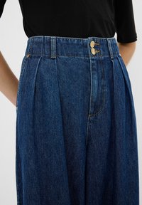 Pantalon en jean bleu foncé taille haute avec plis, avec deux boutons dorés et une ceinture, porté avec un haut noir.