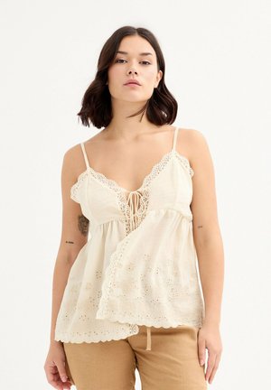 Top sin mangas de color beige claro con un borde de encaje, patrones florales bordados y un detalle atado en el frente, combinado con pantalones marrón claro.