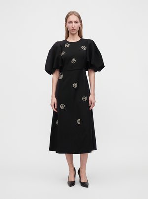 Donna che indossa un vestito midi nero con maniche a sbuffo e motivi floreali decorativi bianchi, abbinato a décolleté nere con punta, in piedi davanti a uno sfondo semplice.