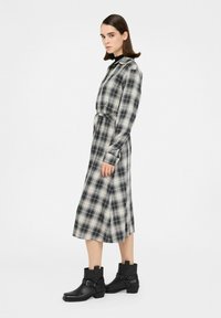 Ung kvinde iført en sort- og hvidternet langærmet kjole og sorte ankelstøvler, stående mod en ensartet lys baggrund.